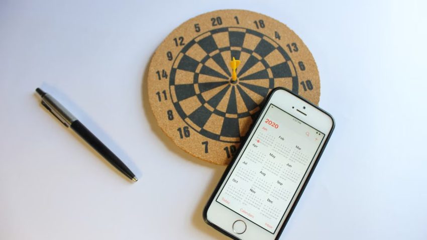 A smartphone with a 2020 calendar next to a dartboard and pen on a white surface.